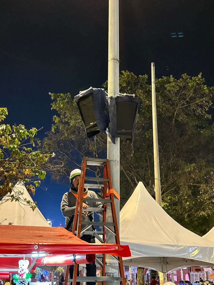 Técnico con casco sobre una escalera, instalando una cabina de sonido en lo alto de un poste, de noche y sobre las carpas de una feria